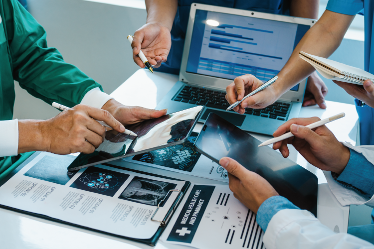 Several people in medical uniforms work together at a table using tablets, a clipboard, and a laptop displaying charts and patient information.
