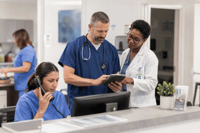 Two healthcare professionals review information on a tablet while a nurse answers the phone at a hospital or clinic reception desk.