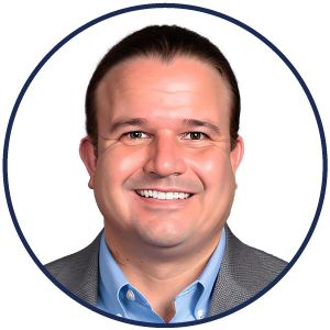 Smiling man with dark, slicked-back hair, wearing a light blue shirt and gray blazer, shown in a circular frame with a white background—evoking the calm confidence of an ambient AI patient experience.