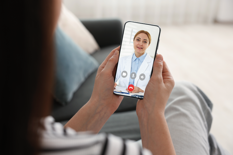 A person holds a smartphone, video chatting with a female doctor who is wearing a white coat and blue shirt. The background shows a sofa, enhancing the ambient AI patient experience by providing comfort and convenience during the virtual consultation.