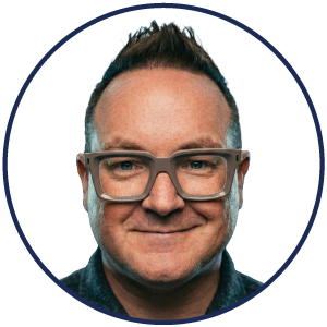 A man with short, dark hair, wearing large, square glasses and a dark shirt, smiles at the camera against a plain white background, embodying the approachable professionalism integral to an exceptional ambient AI patient experience.