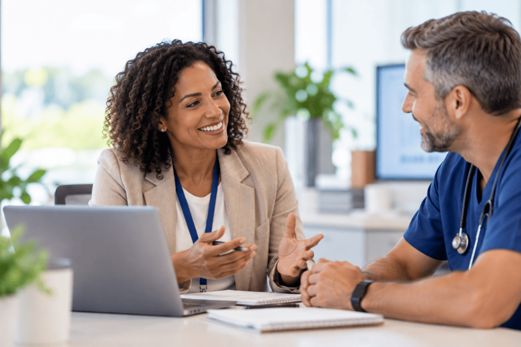 A smiling woman in a business blazer talks to a doctor in scrubs across a desk with a laptop.